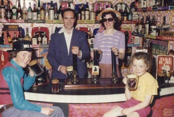 Awkward family photo of four people at a vintage home bar, dressed casually, holding mugs of beer and smiling.