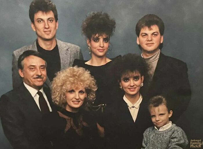 Awkward family photo featuring seven people in '80s style clothing with big hairstyles and serious expressions.