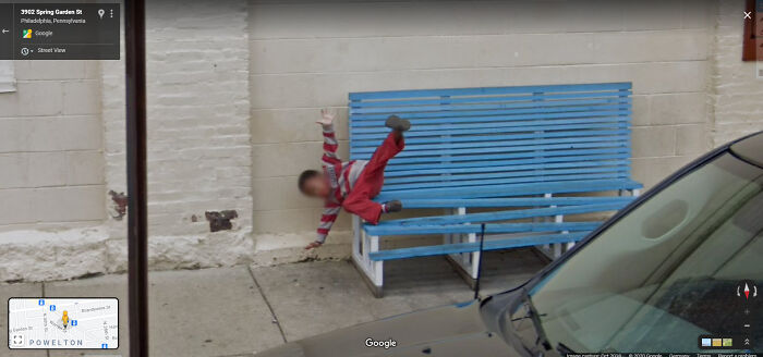 Person in red and white outfit humorously falling off a blue bench, captured on Google Street View.