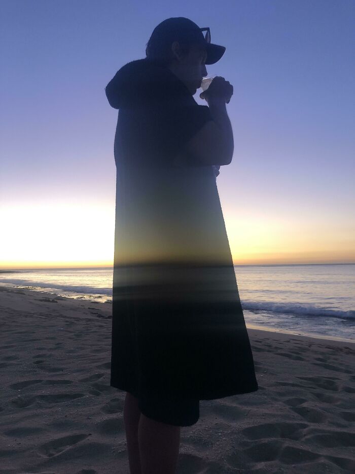 Person silhouetted at sunset on a beach, drinking, creating an interesting, unedited photo effect.