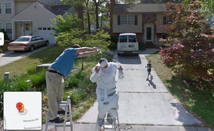 Two people on ladders in a funny scene captured by Google Street View, in a suburban neighborhood.