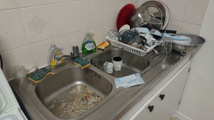 Messy kitchen sink with dirty dishes, overflowing dish rack, illustrating weaponized incompetence.