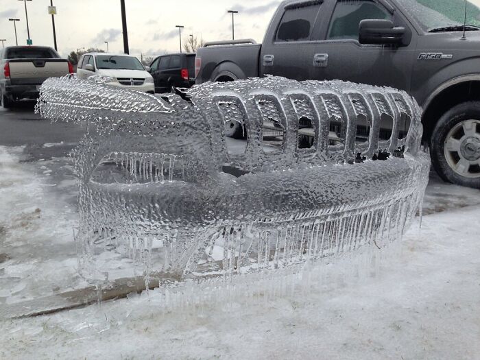 Truck grille covered in ice, creating a unique sculpture in a parking lot during ice-cold winter conditions.