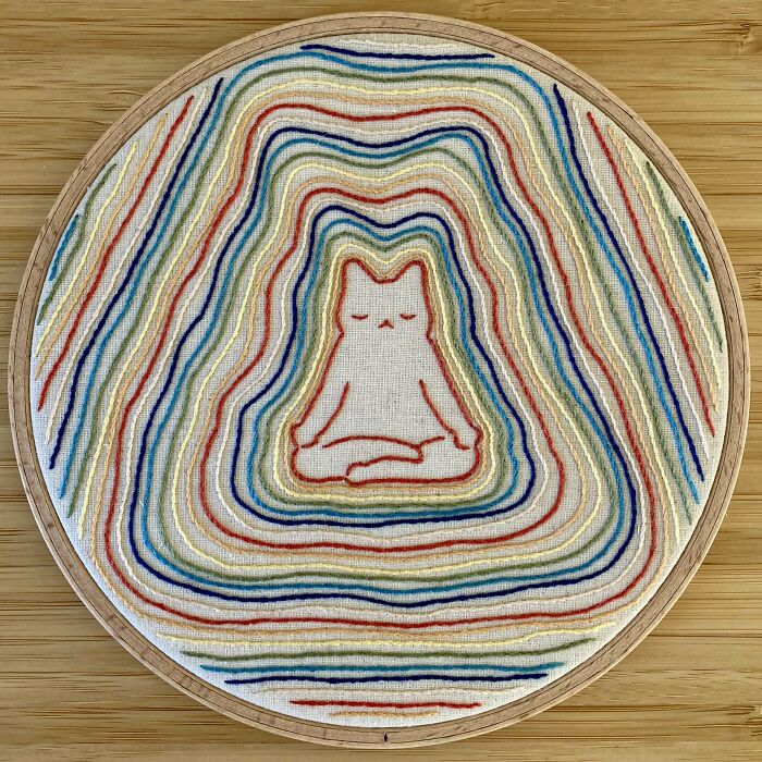 Embroidery design of a meditating cat surrounded by colorful stitched lines in an embroidery hoop on wooden surface.