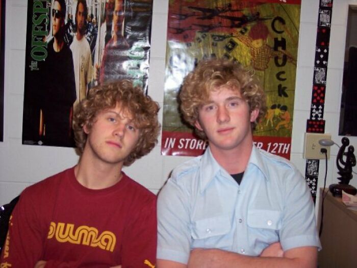 Two men with curly hair resembling uncanny doppelgangers, posing in front of posters.