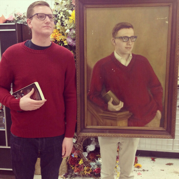 Man in red sweater stands next to a painting with an uncanny doppelganger resemblance, both holding a book.