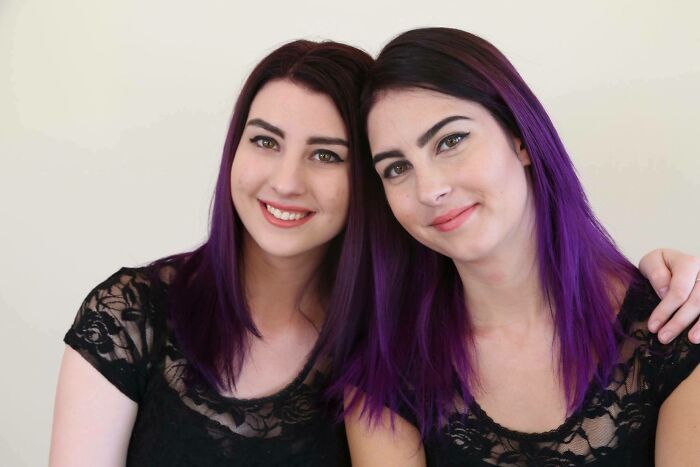 Two women with purple hair and black lace tops, resembling uncanny doppelgangers, smiling side by side.