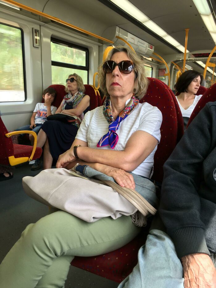 Woman on train wearing sunglasses and a scarf beside another woman with similar appearance; uncanny doppelgangers.