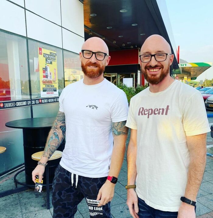 Two men who appear as uncanny doppelgangers standing outside a café, both wearing glasses and casual T-shirts.