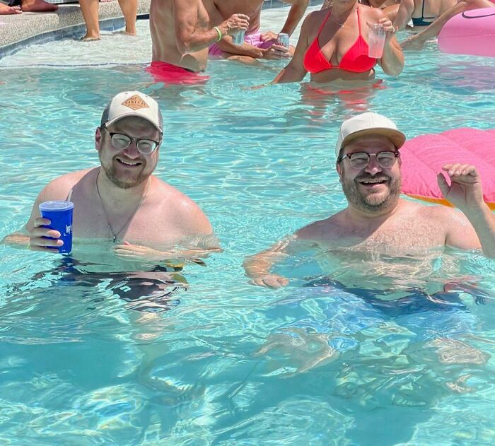 Two uncanny doppelgangers in a pool wearing glasses and hats, holding drinks and smiling.