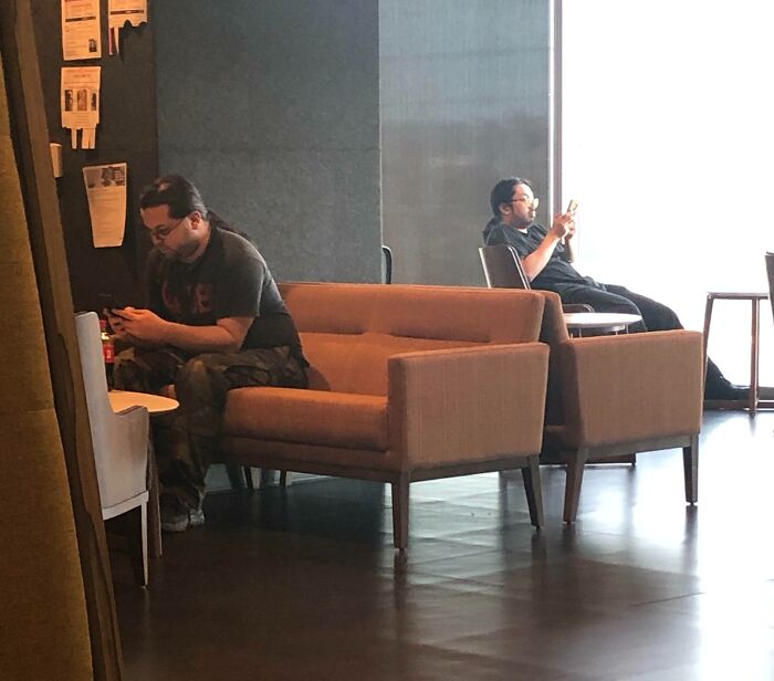 Two men resembling each other, sitting apart in a lounge, both focused on their phones, creating a doppelganger effect.