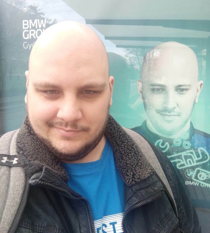 Bald man standing by a poster with his uncanny doppelganger near BMW Group sign, illustrating a Matrix glitch.