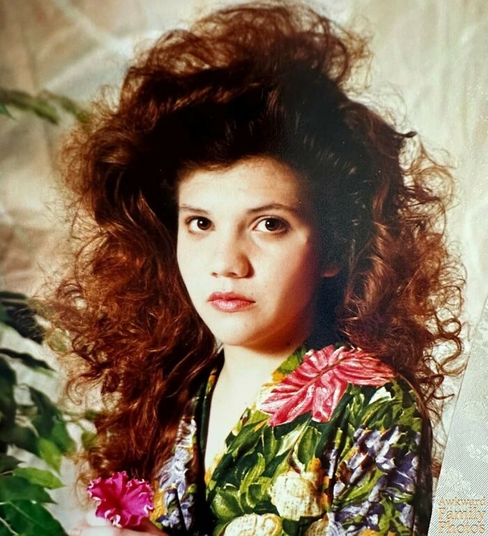 Awkward family photo of woman with voluminous curly hair and floral dress, captured against a soft background.
