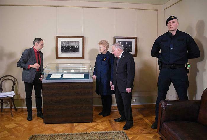 Group of people in a room observing an exhibit, featuring an absolute unit of a security guard standing watch.