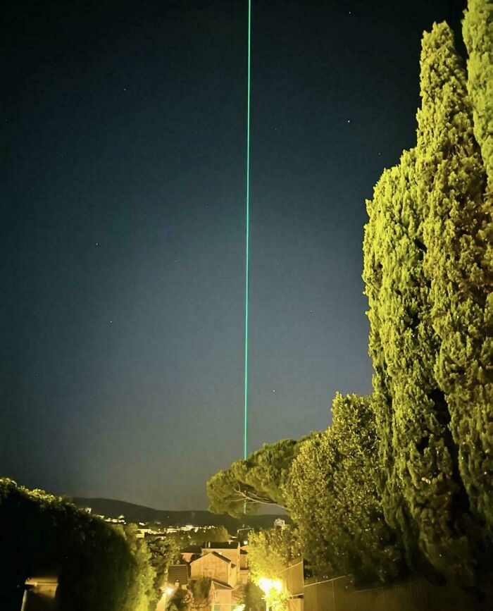 A green laser beam stretching straight into the night sky over a row of trees, showcasing an interesting, not-photoshopped scene.