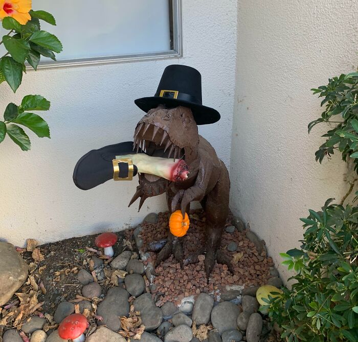 Thanksgiving home decor featuring a T-rex statue with a pilgrim hat and turkey leg, surrounded by autumn foliage.