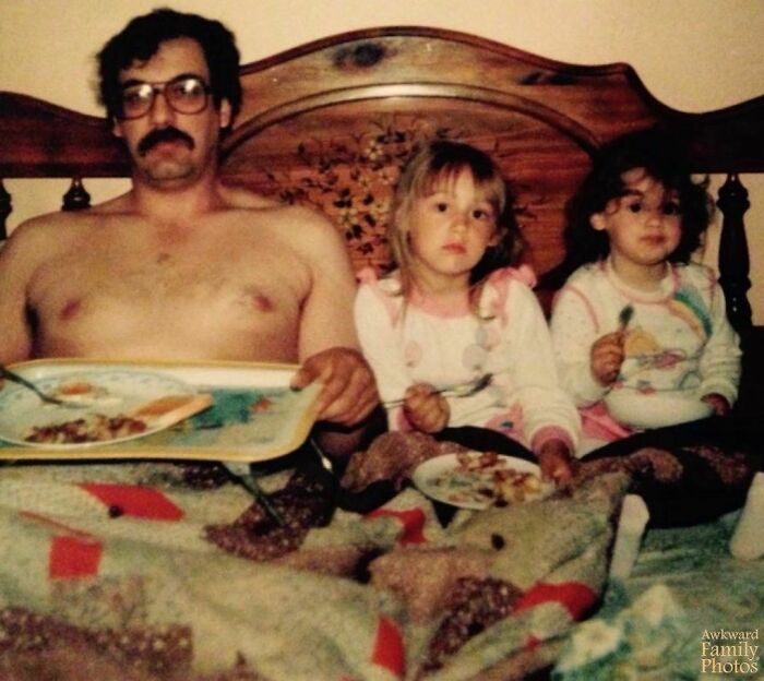 A dad with glasses and two kids in pajamas having an awkward family photo moment while eating in bed.