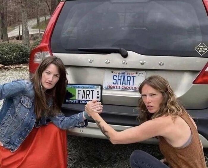 Two women posing with humorous license plates reading "FART" and "SHART" on the back of a Volvo, showcasing a funny moment.