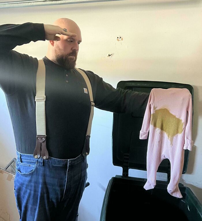 Man showing funny dad moment, holding stained baby clothes near a trash bin in a wholesome dad doing their best scene.