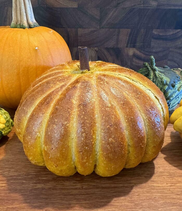 A pumpkin-shaped bread loaf displayed on a wooden counter alongside a real pumpkin, symbolizing a wholesome Thanksgiving.