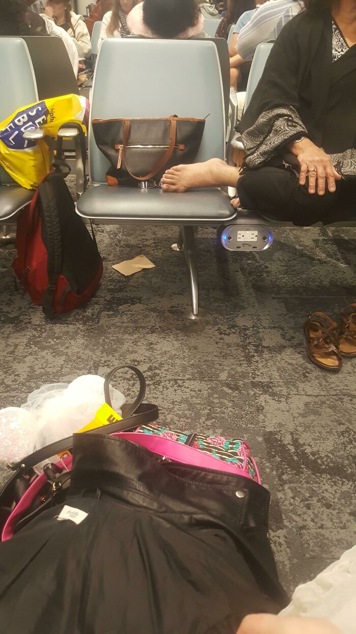 Annoying plane passengers with bare feet on airport seat, surrounded by bags and personal items in a waiting area.