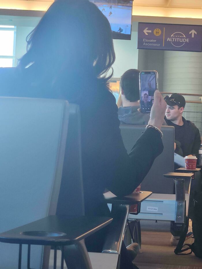 Woman holding phone in airport seating area, possibly capturing annoying plane passengers, with others nearby.