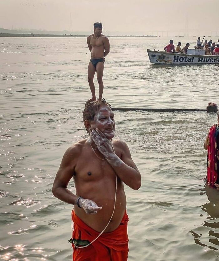 Two men in a fascinating, unedited photo: one stands in water applying soap, another balancing on his head by the river.