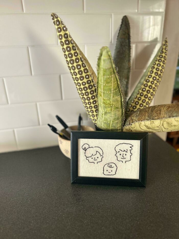 Framed embroidery design featuring simple stitched faces of a family, displayed on a kitchen counter near fabric leaves.