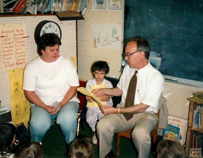 Teacher reading to child and woman in classroom; classic awkward family photo scene.