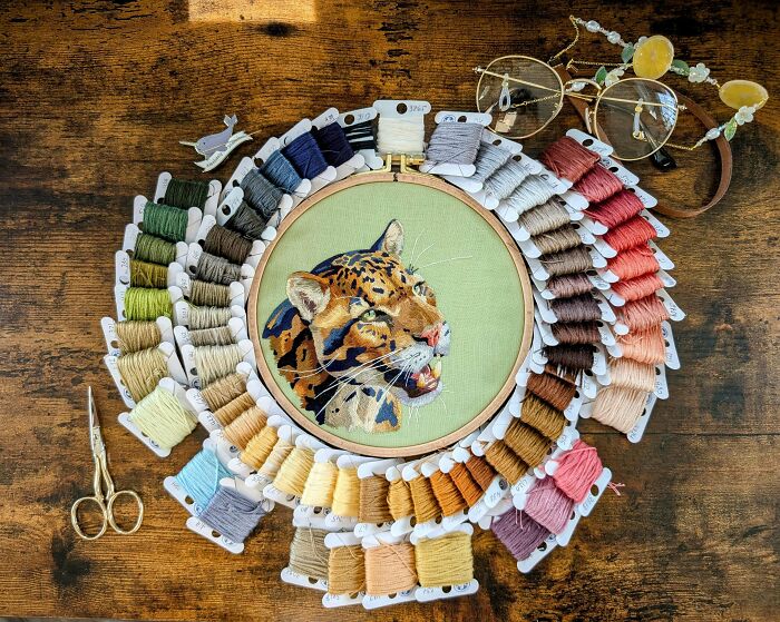 Intricate embroidery design of a leopard surrounded by colorful thread spools, scissors, and glasses on a wooden table.