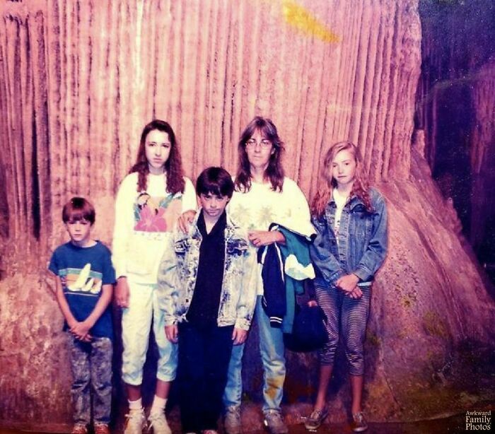 Awkward family photo with five people posing in front of a large tree trunk, wearing 80s style clothes.
