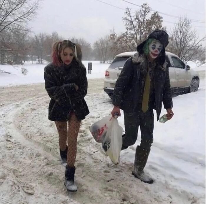 Couple in costume walk through snow, one with a painted face holding a bag.