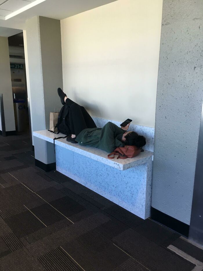 Passenger lying on airport bench with legs up, appearing relaxed and preoccupied with a phone, embodying annoying plane passengers.