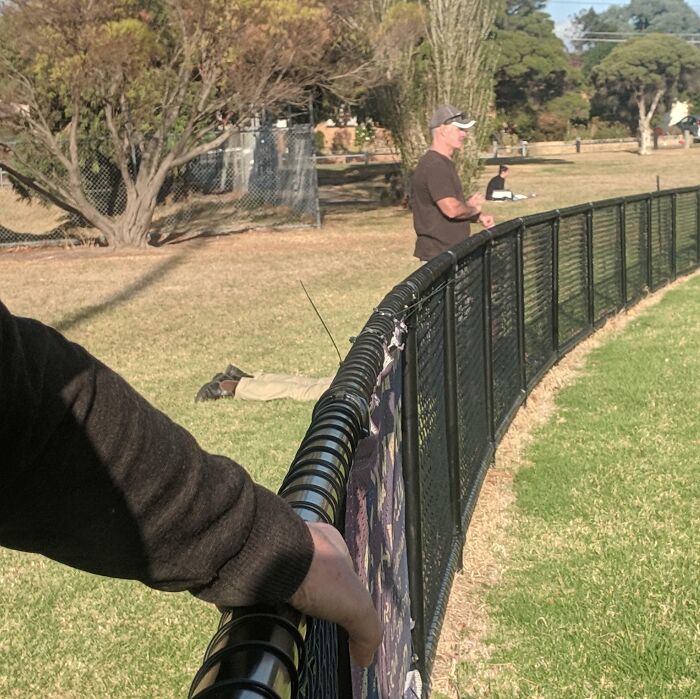 A man stands by a fence, creating an interesting perspective optical illusion, with someone seemingly levitating behind.