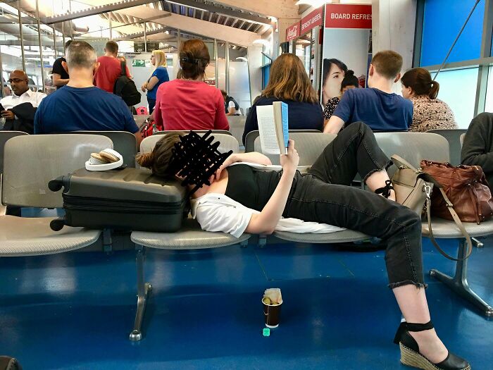 Passenger sprawled across airport seats, reading a book, with luggage as a pillow, illustrating annoying plane passengers.