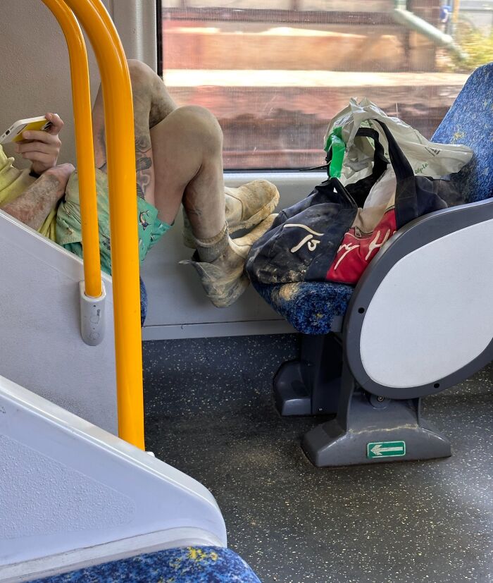 A passenger with dirty boots occupies multiple seats on public transport, bags beside them.