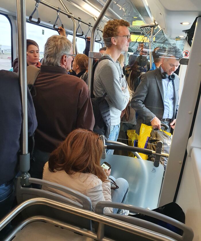 Crowded public transport with passengers standing and a seated woman using her phone.