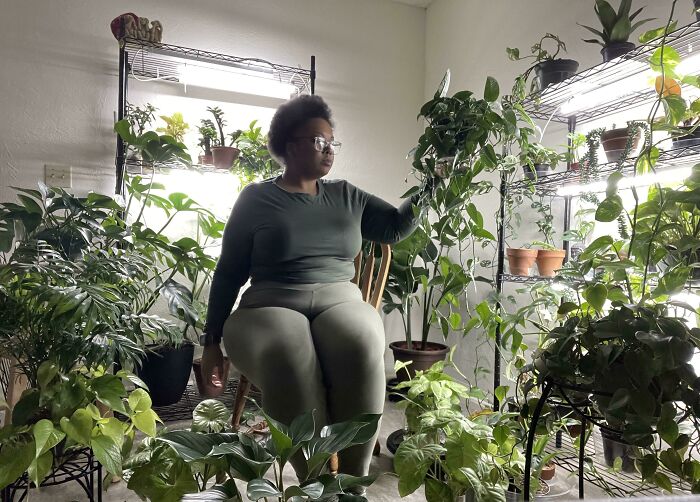 Woman surrounded by lush indoor garden plants, sitting on a chair, tending to greenery on shelves.