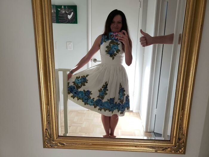 Woman in floral dress takes a mirror selfie, showcasing her outfit with a thumbs-up gesture nearby.