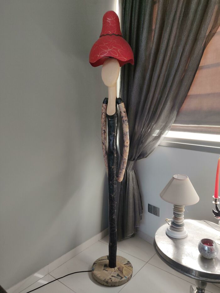 A quirky lamp resembling a person, with a red hat-like shade, stands beside a curtain in a home setting.