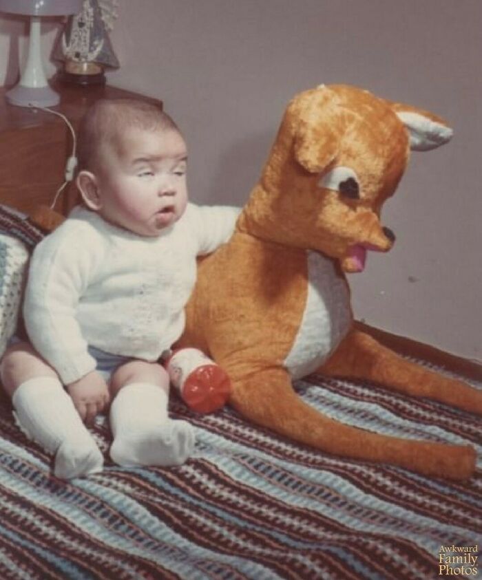Baby with a puzzled look sitting beside a large stuffed reindeer, classic awkward family photo moment.