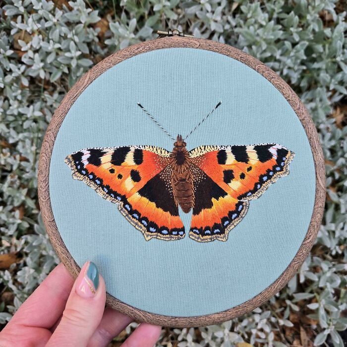 Close-up of a colorful butterfly embroidery design on blue fabric held in a wooden hoop, showcasing beautiful embroidery details.