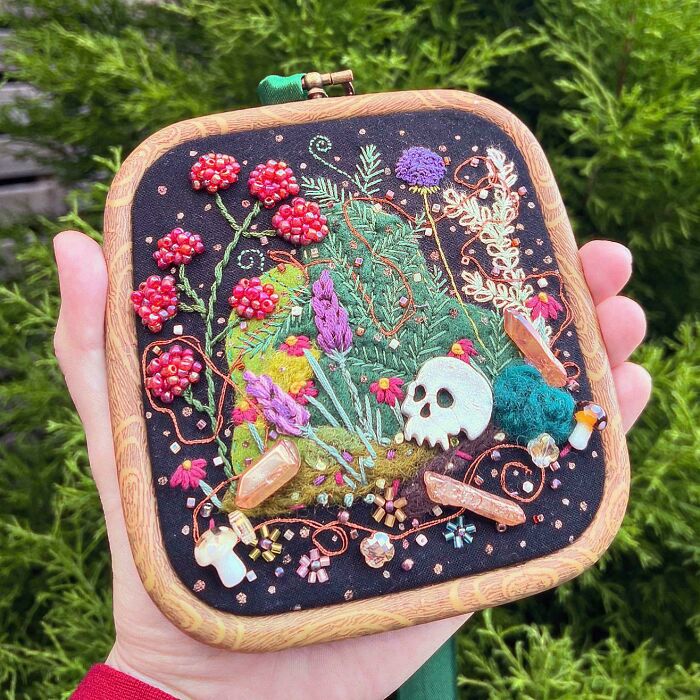 Hand holding a detailed embroidery design featuring flowers, leaves, a skull, and decorative beads on black fabric.