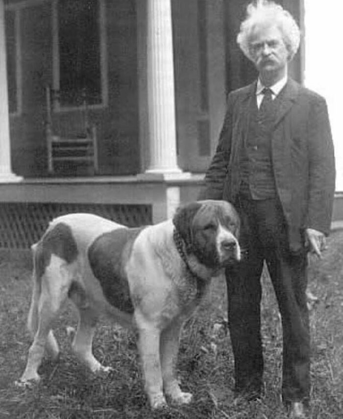 A man in vintage attire stands beside a large, loyal dog in front of a porch, showcasing the timeless bond between pets and family.