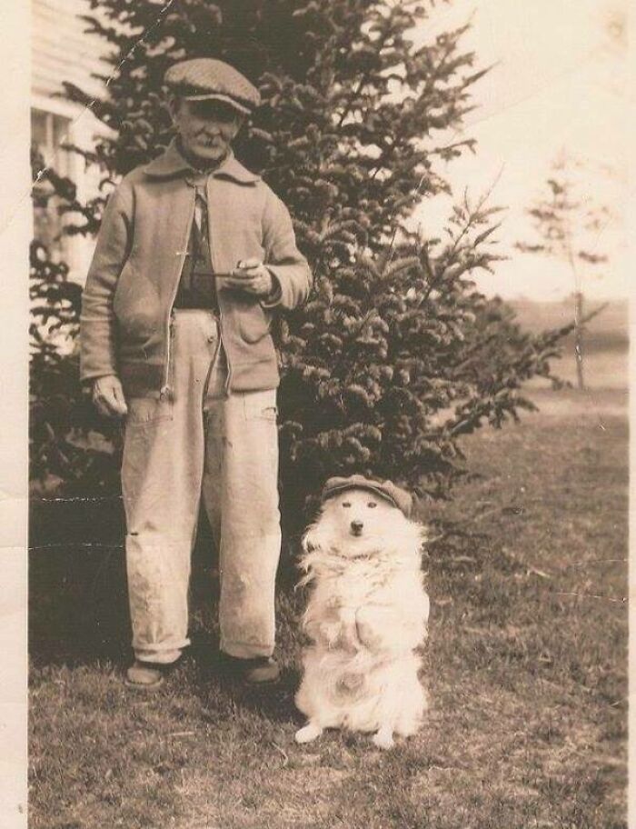 Vintage photo of a man and his dog, both wearing hats, standing in front of a tree, highlighting pets as family.