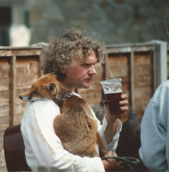 Man with curly hair holds a dog and a pint, showing pets as family in a vintage setting.