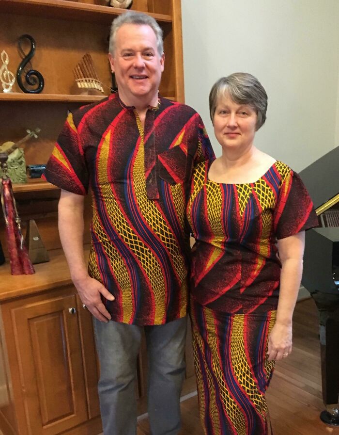 Couple in matching colorful attire stands together in a living room; Amazing-Mothers-In-Law theme.