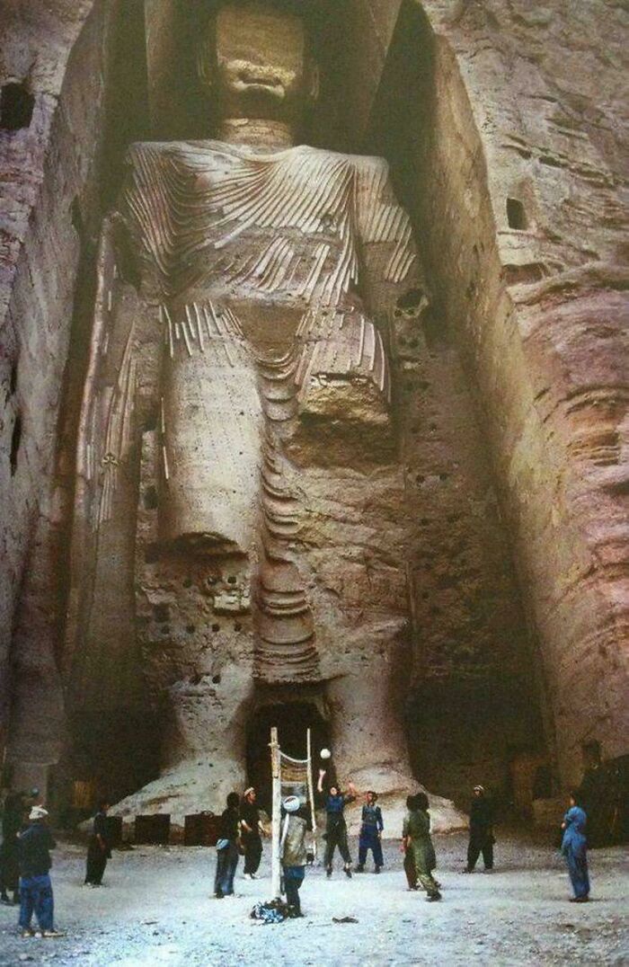 Historical photo of people playing volleyball near a giant ancient statue carved into a rock face.