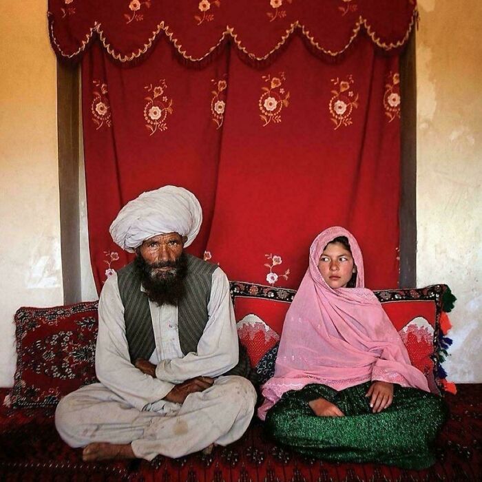 Man and girl in traditional Afghan attire sitting on a patterned sofa, with a red decorative background.