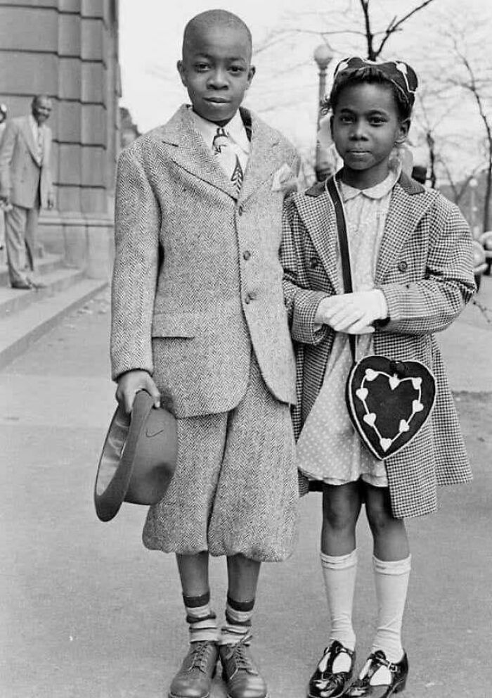 Two children in vintage attire from history, standing on a sidewalk, providing a glimpse into past fashion and culture.